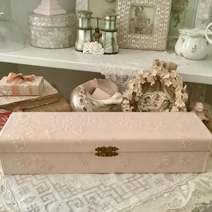 Könnte beinhalten: Eine hellrosa rechteckige Schatulle mit floralem Prägemuster und Messingverschluss. Die Schatulle ist der Mittelpunkt, umgeben von Vintage-Artikeln wie Büchern, einer kleinen Geschenkbox mit Schleife und dekorativen Gläsern.