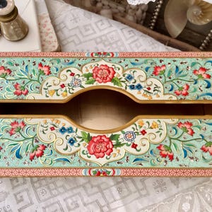 May include: Rectangular tissue box with a floral design. The box features a light teal background with red, pink, blue, and yellow flowers. The edges are trimmed with a pink border. The tissue opening is gold-colored.