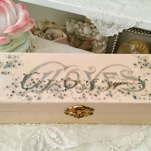 Könnte beinhalten: Eine antike weiße Handschuhbox mit floralem Design und dem Wort "Gloves" in Kursivschrift auf dem Deckel. Die Box hat einen goldenen Verschluss.