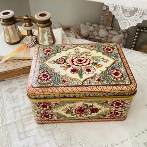 Peut inclure: Une boîte en métal rectangulaire de style vintage avec un motif floral. La boîte présente un fond crème avec des motifs floraux roses, rouges et bleus. Les bords sont dorés. Des jumelles d'opéra et des livres sont en arrière-plan.
