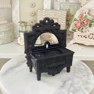 May include: A miniature black vanity with a mirror, ornate detailing, and four legs. The vanity is made of a dark material and sits on a white marble surface. The vanity is a decorative item.