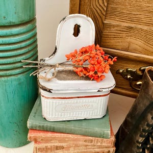 May include: A white vintage match holder with red trim and the word "Allumettes" is displayed on a stack of old books. The holder is filled with orange berries and sits next to a turquoise container and a wooden cabinet.