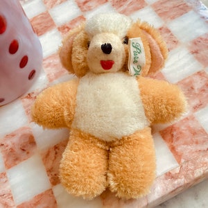 Puede incluir: Un peque&ntilde;o oso de peluche de color crema con brazos y piernas de piel marr&oacute;n. El oso tiene una boca roja y ojos negros. Lleva una etiqueta verde que dice "Pura Lana". El oso est&aacute; sentado sobre una superficie a cuadros rosa y blanca.