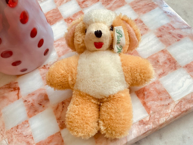 Puede incluir: Un peque&ntilde;o oso de peluche de color crema con brazos y piernas de piel marr&oacute;n. El oso tiene una boca roja y ojos negros. Lleva una etiqueta verde que dice "Pura Lana". El oso est&aacute; sentado sobre una superficie a cuadros rosa y blanca.