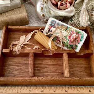 May include: A vintage wooden tray with three compartments, holding a decorative card with floral imagery, a bundle of candles, and dried raffia. The tray is made of stained wood and has a rustic appearance.
