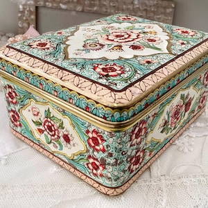Peut inclure: Une boîte en étain décorative vintage avec un motif floral. La boîte est peinte avec un fond blanc et présente un motif floral coloré dans des tons de rose, rouge, vert et bleu. La boîte a une bordure dorée autour des bords.