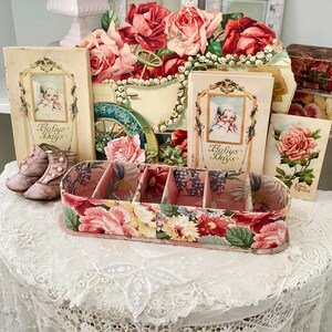 May include: A vintage floral-patterned box with four compartments, surrounded by antique books and floral decorations. The books have the title "Baby's Days" on the cover.