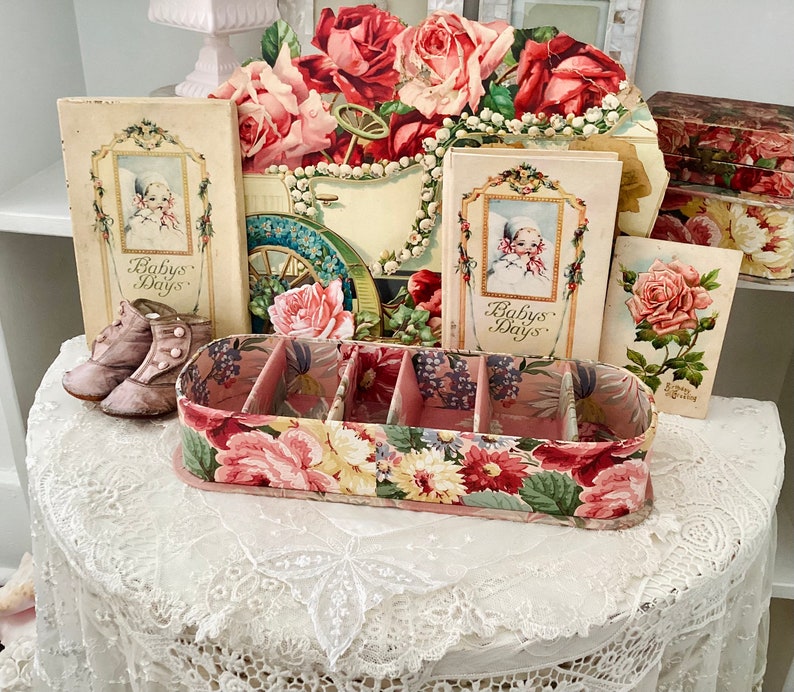 May include: A vintage floral-patterned box with four compartments, surrounded by antique books and floral decorations. The books have the title "Baby's Days" on the cover.