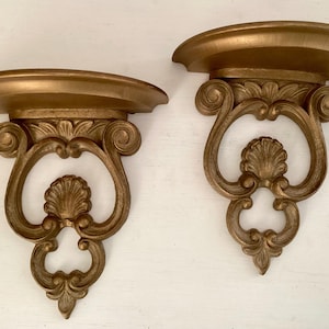 May include: Two ornate, gold-colored wall shelves. Each shelf has a rounded top and a decorative, scrolled bracket with a shell-like design in the center. The shelves are made of a solid material and are designed for displaying items.