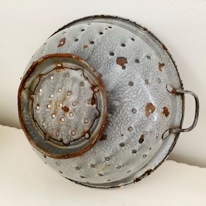 Könnte beinhalten: Vintage graue und weiße Emaille-Küchensieb mit einem kleineren Sieb darin. Das Küchensieb hat einen Griff und zahlreiche kleine Löcher. Das Emaille weist Alterserscheinungen mit Rostflecken und Abnutzung auf, was ihm ein rustikales Aussehen verleiht.