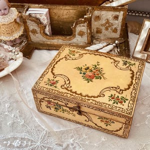 May include: A small, ornate wooden box with a hinged lid. The box is decorated with a floral pattern in shades of pink, yellow, and green. The box is closed and sitting on a white lace tablecloth.