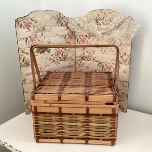 May include: A rectangular, woven wicker picnic basket with a handle. The basket is light brown with darker brown accents. Behind the basket is a floral patterned headboard with a lace trim.