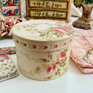 May include: A circular vintage box with a lid, decorated with pink rose floral patterns and green leaves. The box has a cream-coloured background with vertical stripes. Other vintage items are in the background.