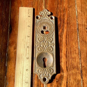 Könnte beinhalten: Verzierte Messing-Türplatte mit Schlüsselloch und dekorativen Schnörkeln. Die Platte ist etwa 18 cm lang und wird mit einem Holzlineal zur Veranschaulichung gezeigt. Die Platte hat oben eine quadratische Öffnung und unten ein Schlüsselloch.