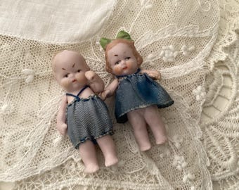 May include: Two porcelain dolls wearing blue denim dresses. The dolls are posed sitting on a white lace doily.