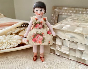 以下が含まれることがあります： 黒い短い髪をした磁器製のドールが、花柄のドレスを着ています。ドールは白い表面に立っており、背景には装飾的な箱があります。
