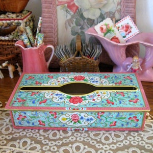 Peut inclure: Un couvre-boîte à mouchoirs de style vintage avec un motif floral dans des tons de rose, bleu et vert. Le couvercle est en métal et possède une bordure dorée. Le boîte à mouchoirs est posée sur une nappe en dentelle.