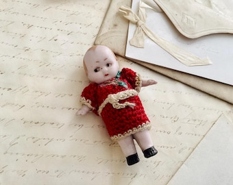 May include: A porcelain doll with painted features wearing a red crocheted dress with white trim. The doll is lying on a sheet of paper with handwritten text.