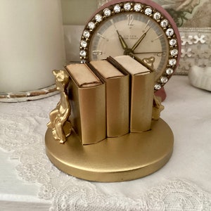May include: Gold-toned decorative bookends with a pair of small, gold-toned figurines sitting on a gold-toned base. The bookends are holding three small, gold-toned books.