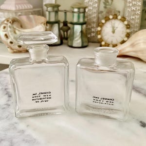 Puede incluir: Dos frascos de perfume cuadrados de vidrio transparente con el texto "CHANEL INC. NEW YORK DISTRIBUTOR SO. J.S.V." impreso en negro. Un frasco tiene un tapón rectangular. Los frascos están sobre una superficie blanca.