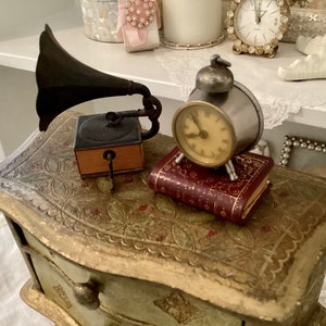 May include: A vintage-style scene featuring a miniature gramophone, a small silver alarm clock, and a tiny red book with gold accents. The items rest on a decorative, aged chest with ornate detailing. The background includes other antique decor.