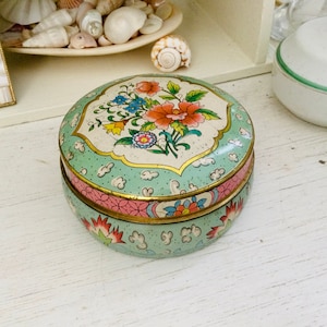 Peut inclure: Une boîte ronde en étain à deux pièces avec un motif floral. Le dessus de la boîte est bleu clair avec une bordure blanche et un motif floral dans des tons de rose, jaune, bleu et vert. Le fond de la boîte est bleu clair avec un motif floral rose et or.
