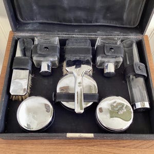May include: An open black leather travel grooming kit with a wooden frame. The kit contains a brush, a razor, two round containers, and three glass bottles with chrome accents. The kit is displayed on a wooden surface.
