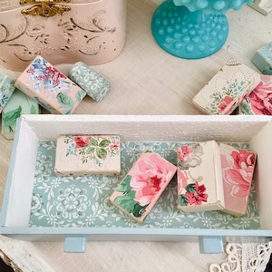 Puede incluir: Un cajón de madera de estilo vintage con un interior azul claro. El cajón está abierto y contiene varias cajas pequeñas con motivos florales en tonos de rosa, blanco y verde.