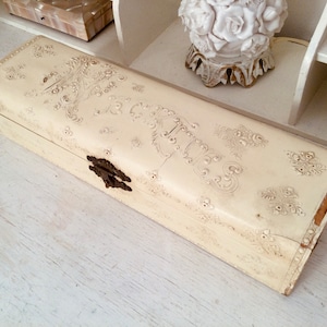 Könnte beinhalten: Eine weiße, rechteckige Schmuckschatulle mit einem verzierten Blumenmuster und einem Messingverschluss. Die Schachtel ist aus Holz und hat einen Vintage-Look.