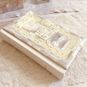 May include: A white book is topped with a rectangular box featuring a mosaic design. The box is made of cream-coloured and iridescent mother-of-pearl tiles, with a floral motif in the centre. The items are on a white lace surface.