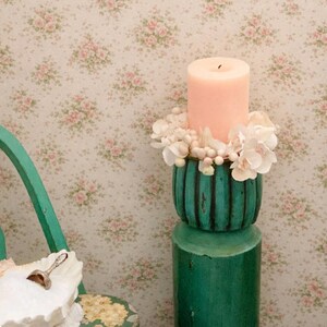 Puede incluir: Una vela de pilar color melocotón se encuentra sobre un portavelas decorativo color turquesa adornado con flores blancas. El portavelas tiene una base cilíndrica y una parte superior acanalada. El fondo presenta un papel pintado floral con rosas rosas.