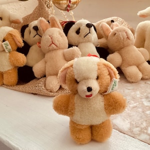 May include: A collection of plush animal toys, including a dog, panda bears, lambs, and rabbits. The dog toy is tan and white with a red mouth and a tag. The other toys are white, tan, and black. The toys are arranged on a white surface.
