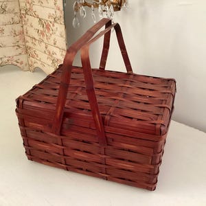 May include: A rectangular, woven bamboo picnic basket with a dark reddish-brown finish. The basket has a hinged lid and a sturdy handle for carrying. The basket is empty and sits on a white surface.