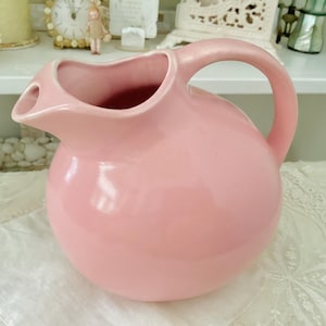 May include: A pink ceramic pitcher with a round body and a curved handle. The pitcher is sitting on a white lace tablecloth.