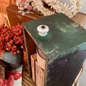 Könnte beinhalten: Eine dunkelgrüne, abgenutzte Holzkiste mit einem weißen Keramikknauf. Die Kiste ist offen und gibt den Blick auf ihr Inneres frei. Die Szene umfasst einen Vintage-Koffer, Blumen und ein kleines gerahmtes Bild, was eine rustikale Ästhetik erzeugt.