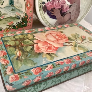 May include: Vintage rectangular tin box with a floral rose design. The box features a light blue background with a large pink rose and smaller roses around the edges. Other decorative tins are visible in the background.