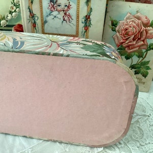 May include: A vintage floral patterned box with a pink lid. The box is made of cardboard and features a floral design with green leaves and pink flowers. The lid is closed and the box is sitting on a white surface.