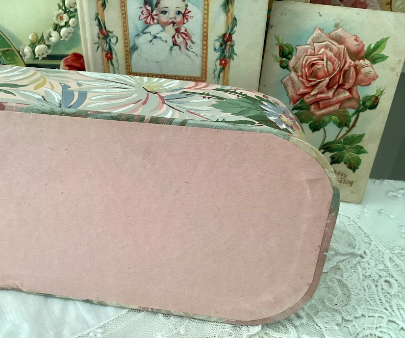 May include: A vintage floral patterned box with a pink lid. The box is made of cardboard and features a floral design with green leaves and pink flowers. The lid is closed and the box is sitting on a white surface.