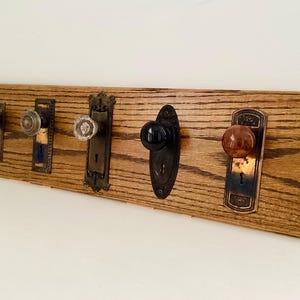 以下が含まれることがあります： 5つのヴィンテージドアノブが付いた木製の壁掛けコートラック。ノブは白、クリア、茶色など様々なスタイルと色があり、ステンドウッドボードに取り付けられています。金具は真鍮と金属の混合です。