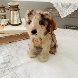 Puede incluir: Un juguete de perro de peluche vintage con pelaje marrón y crema, que lleva una pequeña etiqueta que dice "Molly". El perro está sentado erguido sobre una superficie blanca. El fondo incluye objetos antiguos.
