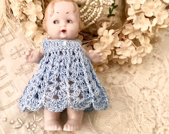 May include: A vintage doll wearing a light blue crocheted dress. The doll has a painted face with rosy cheeks and red lips. The dress is intricately crocheted with a delicate openwork pattern. The doll is posed against a backdrop of lace and cream-colored flowers.