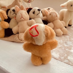 May include: A collection of plush animal toys, including a dog, panda, rabbit, and lamb. The dog toy is light brown with cream-colored accents and a red ribbon. Other toys are white and cream-colored.