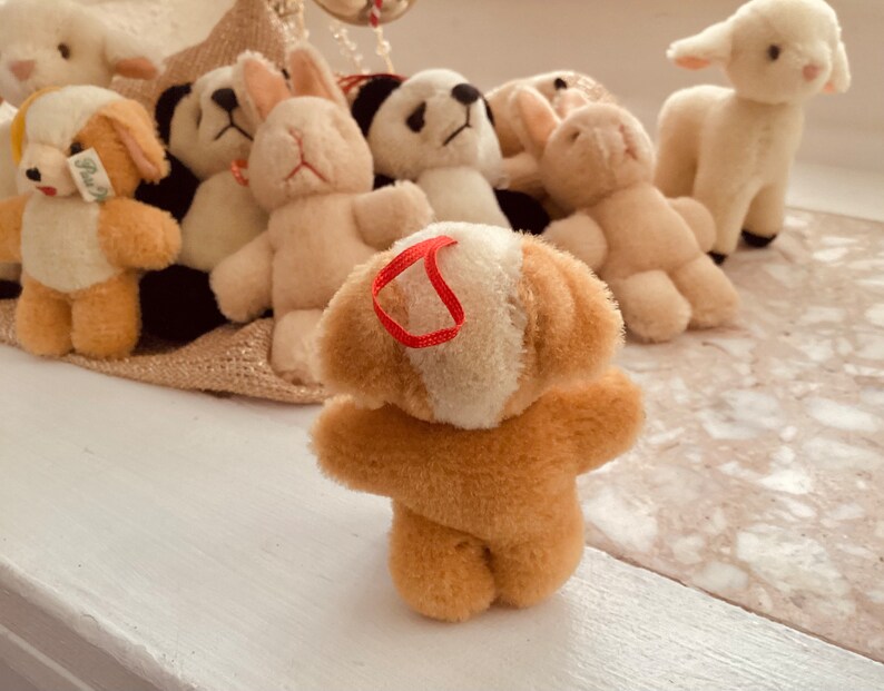 May include: A collection of plush animal toys, including a dog, panda, rabbit, and lamb. The dog toy is light brown with cream-colored accents and a red ribbon. Other toys are white and cream-colored.