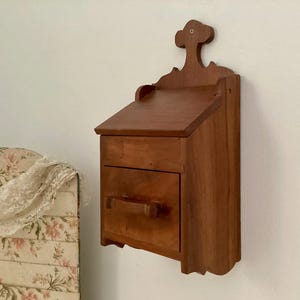 May include: A wooden wall-mounted cabinet with a drawer and a decorative top. The cabinet is made of a warm-toned wood and is mounted on a white wall. The drawer has a simple, rectangular handle.