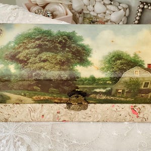 Könnte beinhalten: Eine antike Holzkiste mit einem Scharnierdeckel und einem Messingverschluss. Die Kiste ist mit einer gemalten Szene einer ländlichen Landschaft mit einem Bauernhaus, Bäumen und einem Teich verziert. Die Kiste ist mit marmoriertem Papier ausgekleidet.