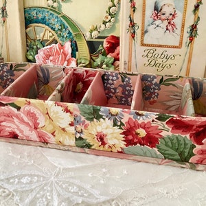 May include: An oval-shaped box with four compartments, featuring a pink and green floral pattern. The box is made of cardboard and has a pink trim. It is resting on a white lace tablecloth.