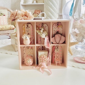 Puede incluir: Una caja expositora rosa claro con múltiples compartimentos llena de objetos decorativos. La caja contiene etiquetas, un pequeño bolso, una jarra en miniatura y otros adornos. Los artículos son en tonos rosa y blanco, con estampados florales y a rayas. El tema general es vintage y femenino.