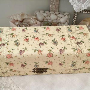 Könnte beinhalten: Eine kleine, rechteckige, cremefarbene Schachtel mit einem Blumenmuster und einem Perlenbesatz. Die Schachtel hat einen Klappdeckel und einen Messingverschluss.