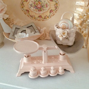 May include: A pale pink vintage-style scale with a round platform and weights. A small basket with a seashell sits on the other side. The scene is decorated with other vintage items, including a floral plate and decorative frames.