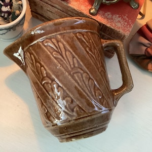 Könnte beinhalten: Ein brauner Keramikkrug mit Henkel und Ausguss. Der Krug hat ein strukturiertes, diagonales Blattmuster. Der Krug ist wahrscheinlich Vintage oder antik und ein dekorativer Gegenstand.